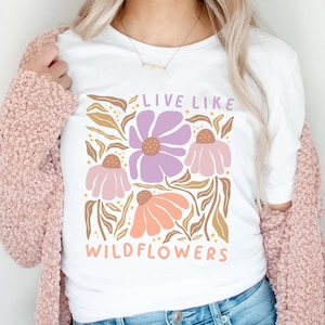May include: White t-shirt with a floral design featuring purple, pink, and orange flowers, and the text "LIVE LIKE WILDFLOWERS". The shirt is paired with blue jeans and a pink cardigan.