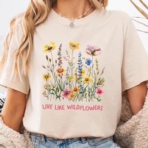 Puede incluir: Una camiseta beige con estampado floral y el texto "Live Like Wildflowers" en rosa.