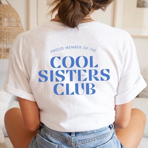 Könnte beinhalten: Ein weißes T-Shirt mit dem Text "PROUD MEMBER OF THE COOL SISTERS CLUB" in Blau. Das T-Shirt wird von einer Person getragen, die Jeansshorts trägt. Das Bild ist von hinten aufgenommen.
