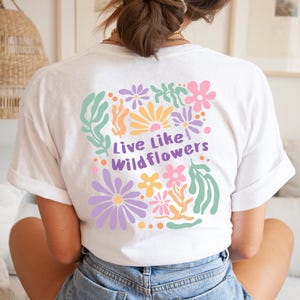 K&ouml;nnte beinhalten: Wei&szlig;es T-Shirt mit einem farbenfrohen Blumendesign und dem Text "Live Like Wildflowers" auf dem R&uuml;cken. Das Design umfasst Blumen in Lila, Rosa, Gelb und Orange, mit gr&uuml;nen Bl&auml;ttern. Das Shirt ist aus weichem Material.