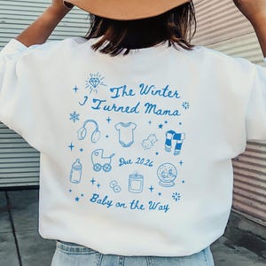 May include: White sweatshirt with blue text and illustrations. The text reads "The Winter I Turned Mama" with baby-themed icons and "Due 2026" and "Baby on the Way". The sweatshirt is a casual style.