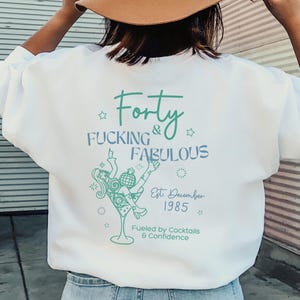 Op de afbeelding: Witte sweatshirt met de tekst "Forty Fucking & Fabulous" in turquoise script. Onder de tekst staat een afbeelding van een vrouw in een cocktailglas, met de tekst "Est. December 1985" en "Fueled by Cocktails & Confidence".