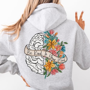 Może przedstawiać: Jasnoszara bluza z kapturem z grafiką na plecach. Grafika przedstawia ilustrację mózgu z kolorowymi kwiatami i napisem "Be Kind to Your Mind" na banerze. Bluza ma kaptur i długie rękawy.