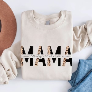 Könnte beinhalten: Ein beiger Pullover mit dem Wort "MAMA" in schwarzen Buchstaben mit Leopardenmuster. Der Text "Emily & Liam" ist darunter in Kursivschrift geschrieben.