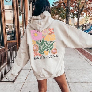 Pode incluir: Um moletom de cor creme com um design floral e o texto "Bloom as you are" nas costas.