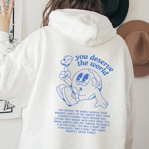 Op de afbeelding: Witte hoodie met een blauwe afbeelding van een cartoon-aarde die een bloem vasthoudt. De tekst "you deserve the world" is in blauw op de achterkant van de hoodie gedrukt.