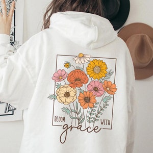 May include: White hoodie with a floral design on the back. The design features a variety of flowers in shades of pink, orange, yellow, and red. The text "Bloom with grace" is printed below the flowers.