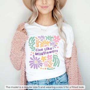 K&ouml;nnte beinhalten: Wei&szlig;es T-Shirt mit einem farbenfrohen Blumendesign und dem Text "Live Like Wildflowers". Das Shirt wird mit einer blauen Jeans und einer rosa Strickjacke kombiniert. Das Model tr&auml;gt Gr&ouml;&szlig;e S f&uuml;r eine figurbetonte Passform.