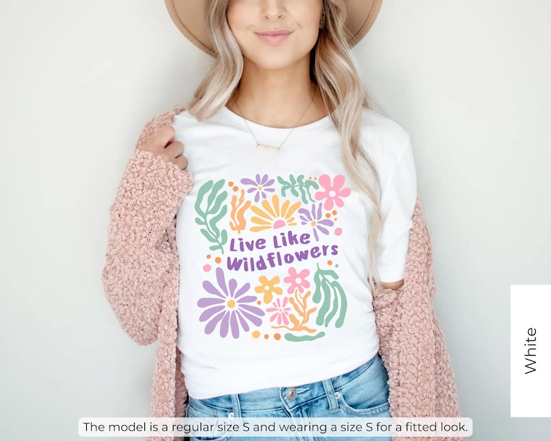 K&ouml;nnte beinhalten: Wei&szlig;es T-Shirt mit einem farbenfrohen Blumendesign und dem Text "Live Like Wildflowers". Das Shirt wird mit einer blauen Jeans und einer rosa Strickjacke kombiniert. Das Model tr&auml;gt Gr&ouml;&szlig;e S f&uuml;r eine figurbetonte Passform.