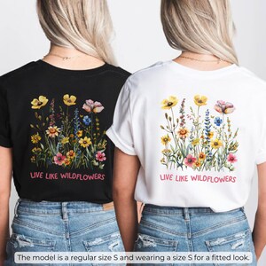 Puede incluir: Dos camisetas con un dise&ntilde;o floral y el texto "Live like wildflowers" impreso en la espalda. La camiseta de la izquierda es negra y la camiseta de la derecha es blanca.