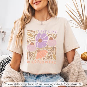 May include: Cream-colored t-shirt with a floral graphic. The design features purple, pink, and orange flowers with the text "LIVE LIKE WILDFLOWERS". The model is wearing a size M for a relaxed fit.