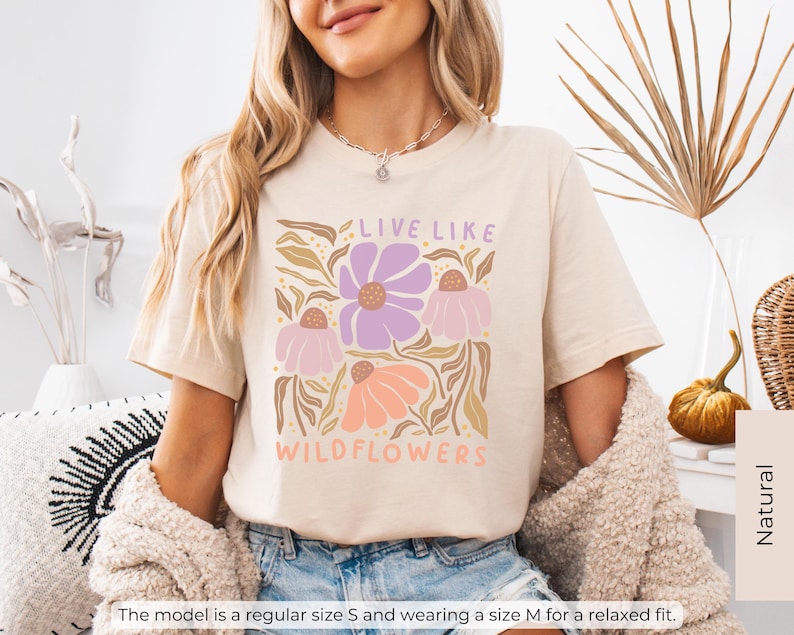 May include: Cream-colored t-shirt with a floral graphic. The design features purple, pink, and orange flowers with the text "LIVE LIKE WILDFLOWERS". The model is wearing a size M for a relaxed fit.