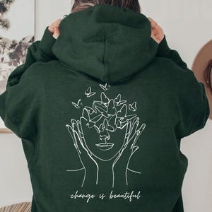 May include: A dark green hoodie with a white line drawing of a face surrounded by butterflies. The text "change is beautiful" is printed below the drawing.