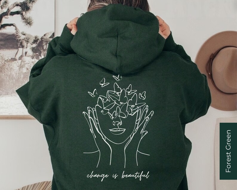 May include: A dark green hoodie with a white line drawing of a face surrounded by butterflies. The text "change is beautiful" is printed below the drawing.