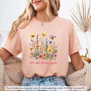 Puede incluir: Una camiseta rosa claro con un dise&ntilde;o floral y el texto "Live Like Wildflowers".
