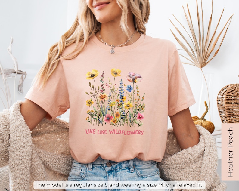 Puede incluir: Una camiseta rosa claro con un dise&ntilde;o floral y el texto "Live Like Wildflowers".