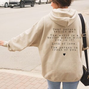 May include: Beige hooded sweatshirt with a black and white text message on the back that reads "Dear person behind me: The world is a better place with you in it! Lots of love. The person in front of you" with a small black heart symbol.