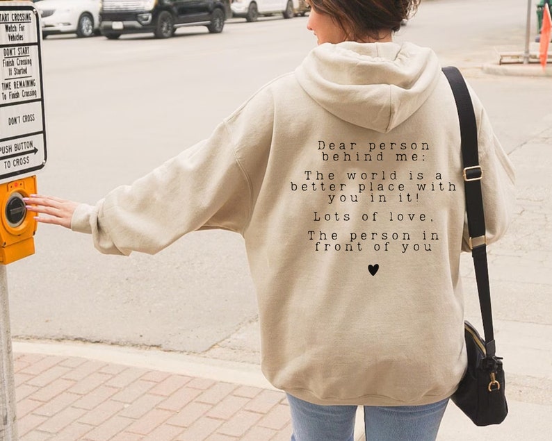 May include: Beige hooded sweatshirt with a black and white text message on the back that reads "Dear person behind me: The world is a better place with you in it! Lots of love. The person in front of you" with a small black heart symbol.