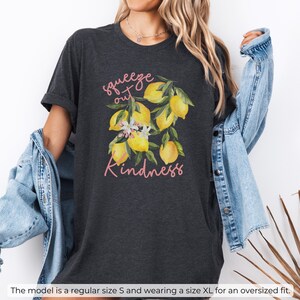 K&ouml;nnte beinhalten: Dunkelgraues T-Shirt aus Heather-Material mit einem Zitronenmotiv und dem Text "squeeze out kindness" in Rosa.
