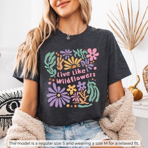 K&ouml;nnte beinhalten: Dunkelgraues meliertes T-Shirt mit einem bunten Blumenmuster und dem Text "Live Like Wildflowers". Das Design umfasst Blumen in Lila, Rosa, Gelb und Gr&uuml;n. Das Model tr&auml;gt Gr&ouml;&szlig;e M f&uuml;r eine lockere Passform.