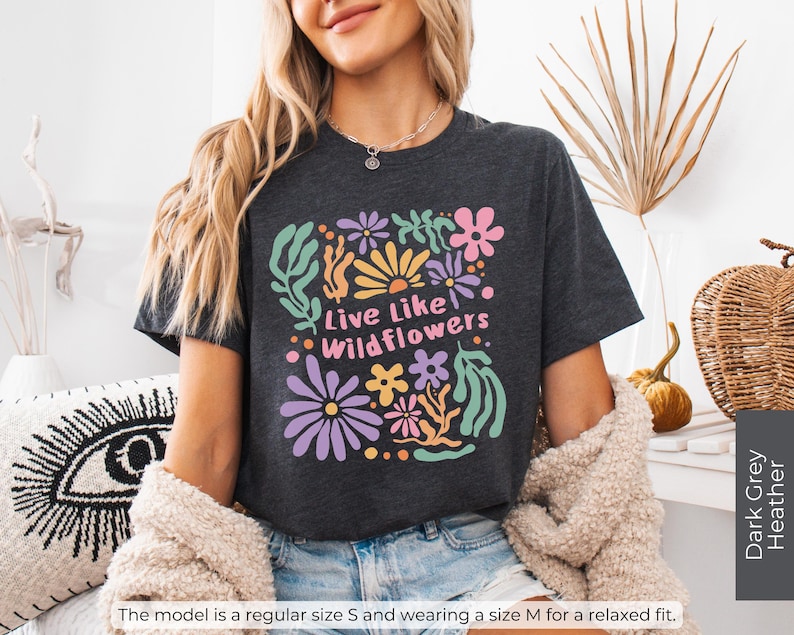 K&ouml;nnte beinhalten: Dunkelgraues meliertes T-Shirt mit einem bunten Blumenmuster und dem Text "Live Like Wildflowers". Das Design umfasst Blumen in Lila, Rosa, Gelb und Gr&uuml;n. Das Model tr&auml;gt Gr&ouml;&szlig;e M f&uuml;r eine lockere Passform.