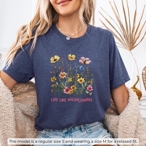 Puede incluir: Una camiseta azul marino con un dise&ntilde;o floral y el texto "LIVE LIKE WILDFLOWERS".