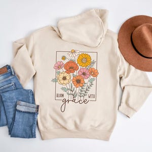 May include: A beige hoodie with a floral design and the text "Bloom with grace" on the front. The hoodie has a hood and long sleeves.