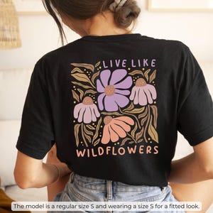 May include: Black t-shirt with a floral design featuring the text "LIVE LIKE WILDFLOWERS." The design includes purple, peach, and tan flowers with green leaves. The shirt is a regular size S and is worn for a fitted look.