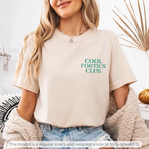 May include: A beige t-shirt with the text "Cool Forties Club" in teal green lettering. The model is wearing a size M for a relaxed fit.