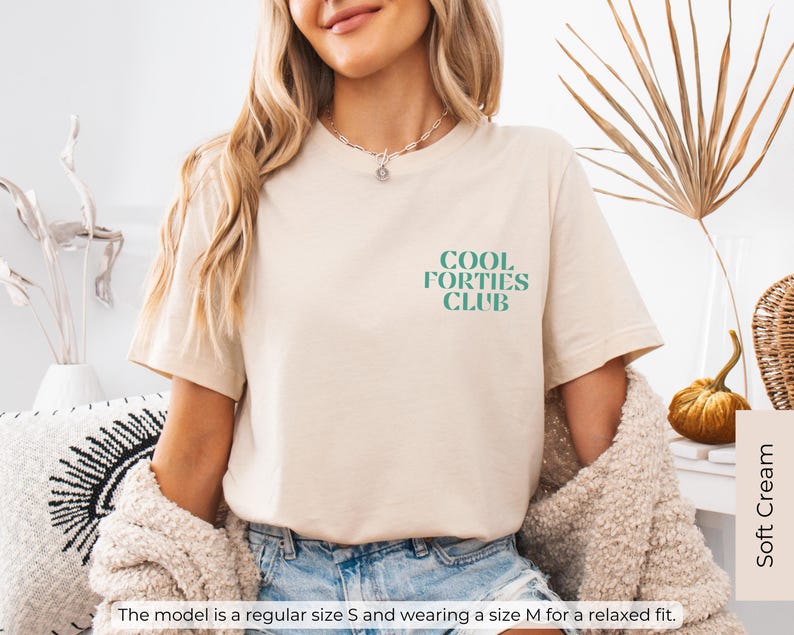 May include: A beige t-shirt with the text "Cool Forties Club" in teal green lettering. The model is wearing a size M for a relaxed fit.