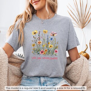 Puede incluir: Camiseta deportiva gris jaspeada con un dise&ntilde;o floral y el texto "Live Like Wildflowers".