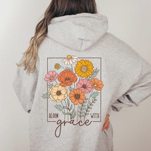 May include: A gray hooded sweatshirt with a floral design on the back. The design features a bouquet of flowers in shades of pink, orange, and yellow, with the text "Bloom with grace" below the flowers.
