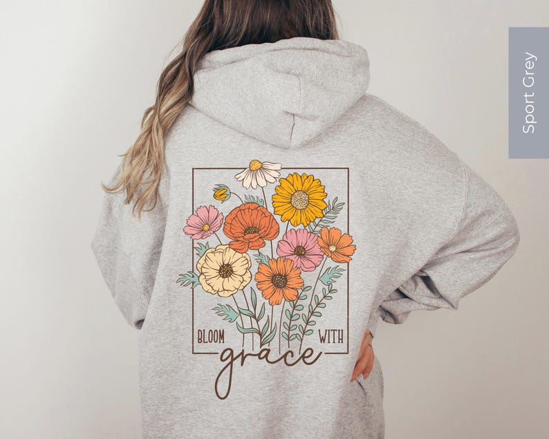 May include: A gray hooded sweatshirt with a floral design on the back. The design features a bouquet of flowers in shades of pink, orange, and yellow, with the text "Bloom with grace" below the flowers.
