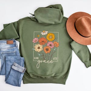 May include: A green hooded sweatshirt with a floral design and the text "Bloom with grace" printed on the front. The sweatshirt is laid flat on a white surface with a pair of blue jeans and a brown hat.