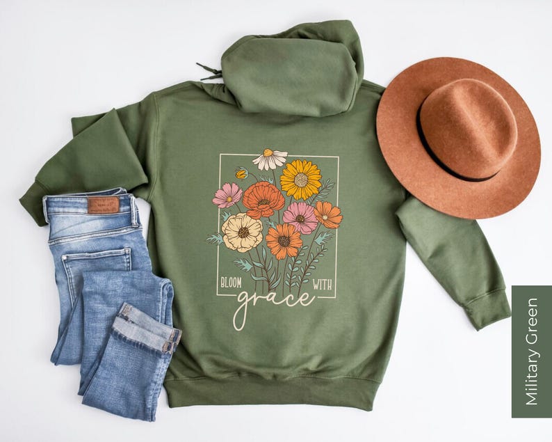 May include: A green hooded sweatshirt with a floral design and the text "Bloom with grace" printed on the front. The sweatshirt is laid flat on a white surface with a pair of blue jeans and a brown hat.