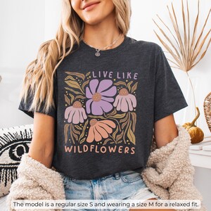 May include: Dark grey heather t-shirt with a floral design and the text "LIVE LIKE WILDFLOWERS" in a whimsical font. The design features purple, pink, and peach flowers with green and gold leaves. The shirt is a relaxed fit.