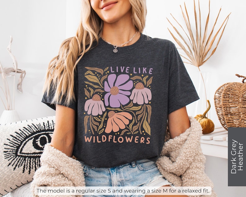 May include: Dark grey heather t-shirt with a floral design and the text "LIVE LIKE WILDFLOWERS" in a whimsical font. The design features purple, pink, and peach flowers with green and gold leaves. The shirt is a relaxed fit.