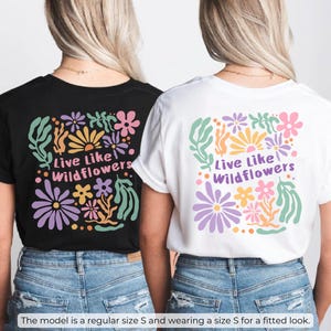 K&ouml;nnte beinhalten: Zwei T-Shirts, eines in Schwarz und eines in Wei&szlig;, mit einem Blumendesign und dem Text "Live Like Wildflowers". Das Design umfasst lila, rosa, gelbe und gr&uuml;ne Blumen und Bl&auml;tter. Die Models tragen die T-Shirts mit blauen Jeans.