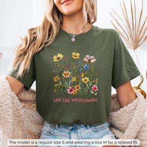 Puede incluir: Una camiseta verde con un dise&ntilde;o floral y el texto "LIVE LIKE WILDFLOWERS" impreso en la parte delantera.