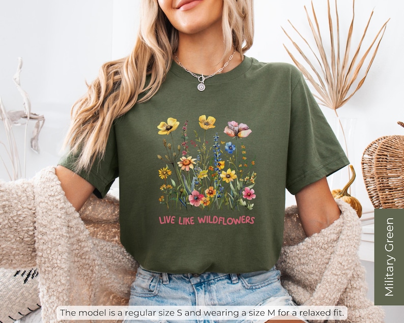 Puede incluir: Una camiseta verde con un dise&ntilde;o floral y el texto "LIVE LIKE WILDFLOWERS" impreso en la parte delantera.