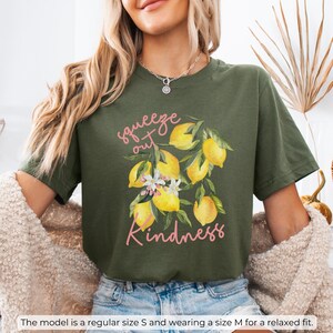 K&ouml;nnte beinhalten: Gr&uuml;nes T-Shirt mit einem Zitronenmotiv und dem Text "squeeze out kindness" in rosa Schrift.