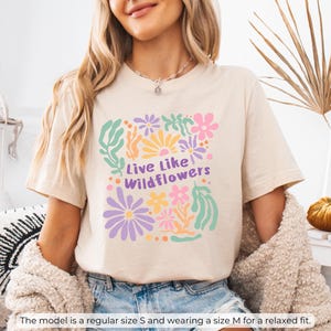 K&ouml;nnte beinhalten: Beiges T-Shirt mit farbenfrohem Blumenmuster und dem Text "Live Like Wildflowers". Das Design umfasst Blumen in Rosa, Lila, Gelb und Gr&uuml;n. Das Shirt hat eine lockere Passform.