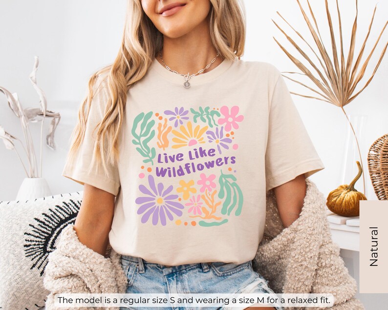 K&ouml;nnte beinhalten: Beiges T-Shirt mit farbenfrohem Blumenmuster und dem Text "Live Like Wildflowers". Das Design umfasst Blumen in Rosa, Lila, Gelb und Gr&uuml;n. Das Shirt hat eine lockere Passform.