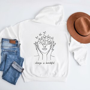 May include: White hooded sweatshirt with a black line drawing of a person with butterflies around their head. The text "change is beautiful" is printed below the drawing.