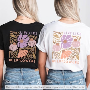 May include: Black and white t-shirts with a floral design. The shirts feature the text "LIVE LIKE WILDFLOWERS" in a stylized font. The floral design includes purple, pink, and orange flowers with brown leaves.