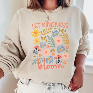 Pode incluir: Uma camisa de moletom bege com um design floral e o texto "Let Kindness Bloom" em letras rosa e laranja.