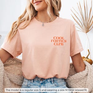 May include: A peach-colored t-shirt with the text "COOL FORTIES CLUB" in orange lettering. The model is wearing a size M for a relaxed fit.