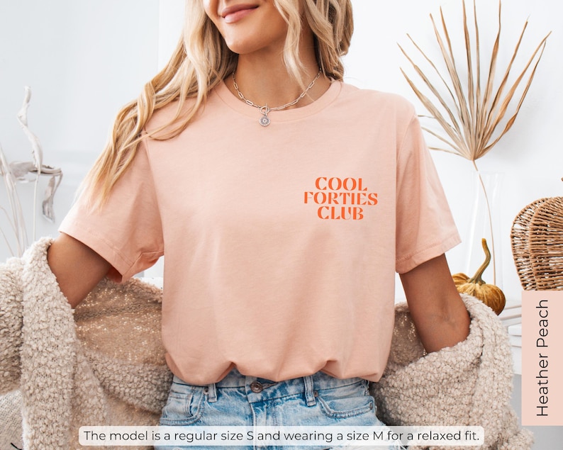 May include: A peach-colored t-shirt with the text "COOL FORTIES CLUB" in orange lettering. The model is wearing a size M for a relaxed fit.