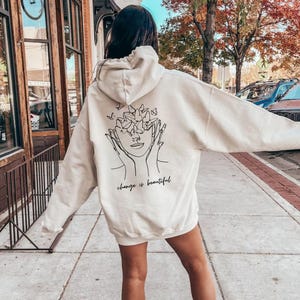 May include: A cream-colored hoodie with a black line drawing of a face with butterflies around it. The text "change is beautiful" is printed below the drawing.