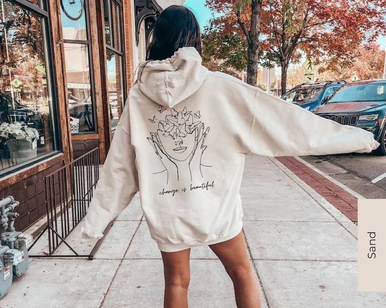 May include: A cream-colored hoodie with a black line drawing of a face with butterflies around it. The text "change is beautiful" is printed below the drawing.